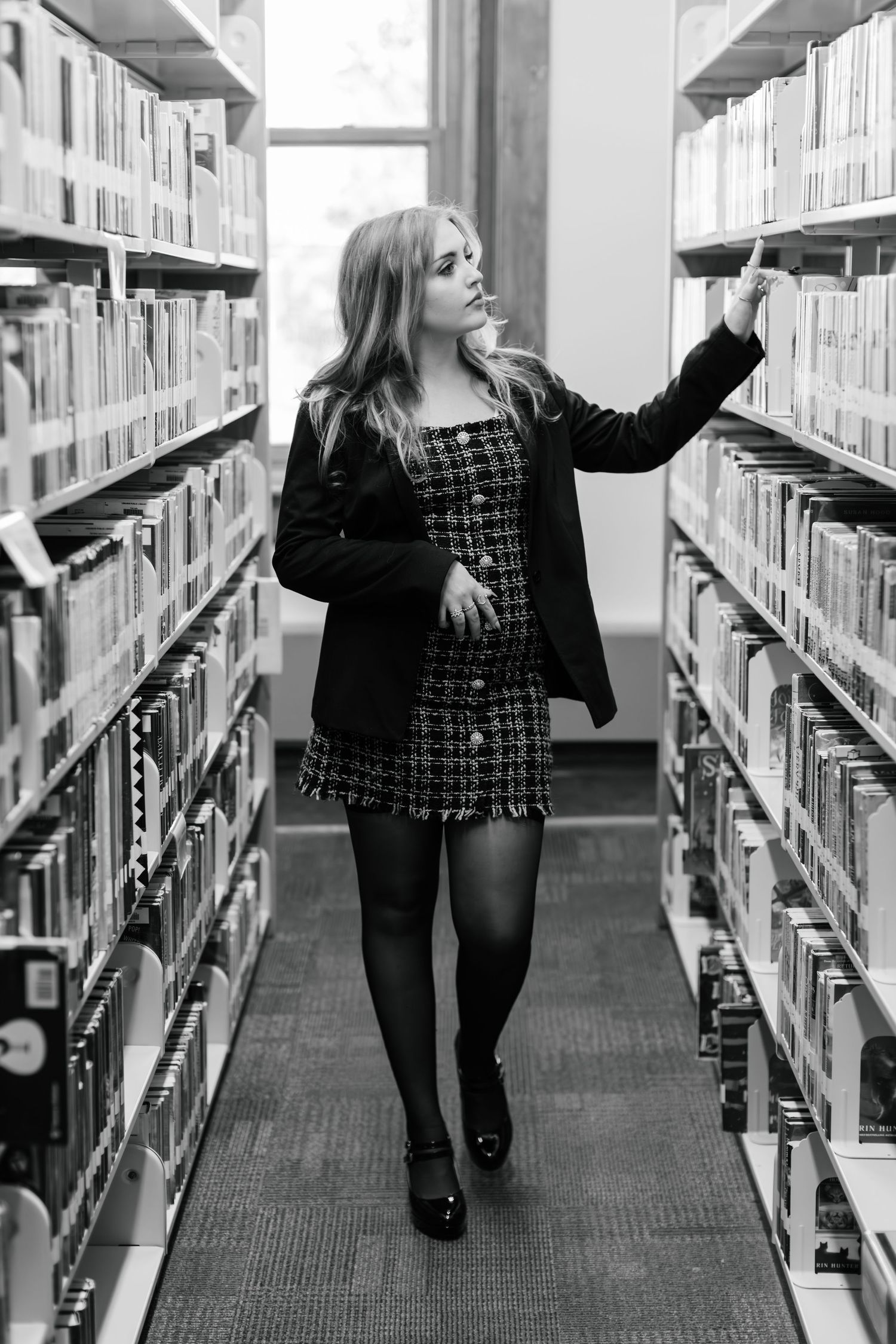 Black and white photo of a person browsing library shelves while wearing a patterned dress and black coat.