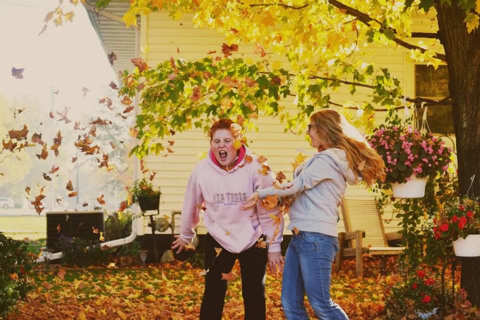 Two people playfully toss autumn leaves in front of a white house under a yellow tree with colorful fall foliage.