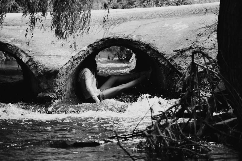 A black and white photo shows someone crouched under a stone bridge arch over a stream.