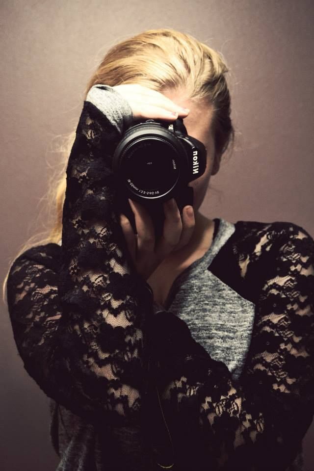 Photographer in black lace sleeves holds a Nikon camera up to take a self portrait in moody lighting.