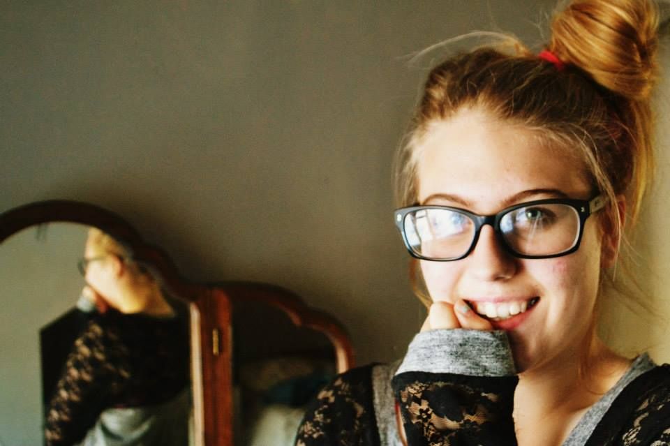 Someone wearing black-rimmed glasses and black lace clothing holds a drink while looking at the camera.