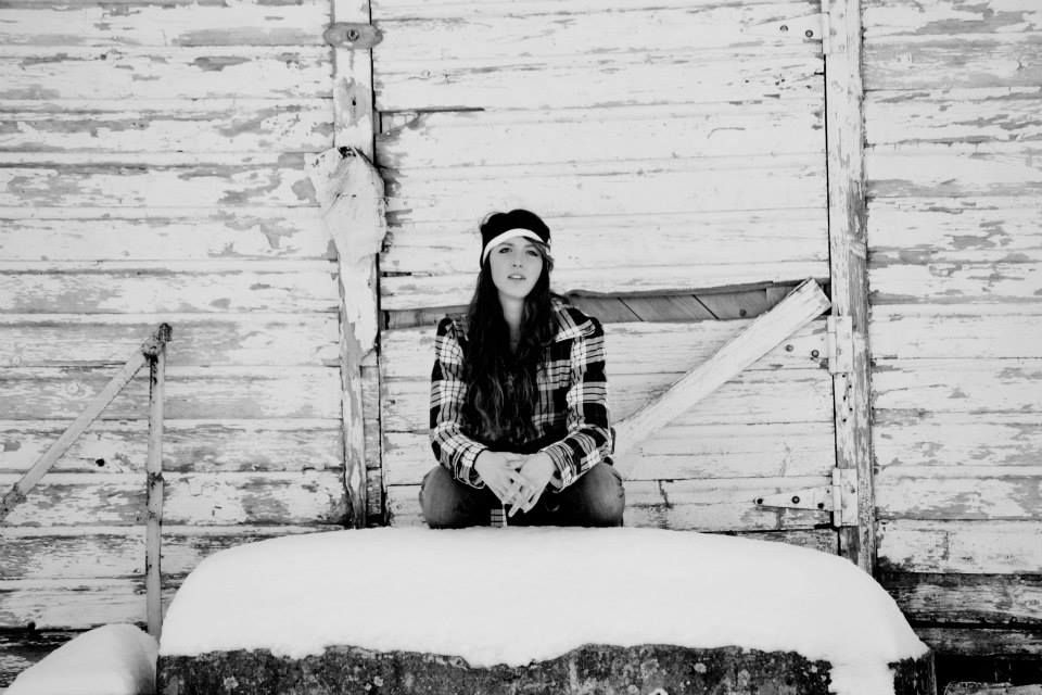 A person in plaid sits on a snow-covered porch step against a rustic wooden wall.