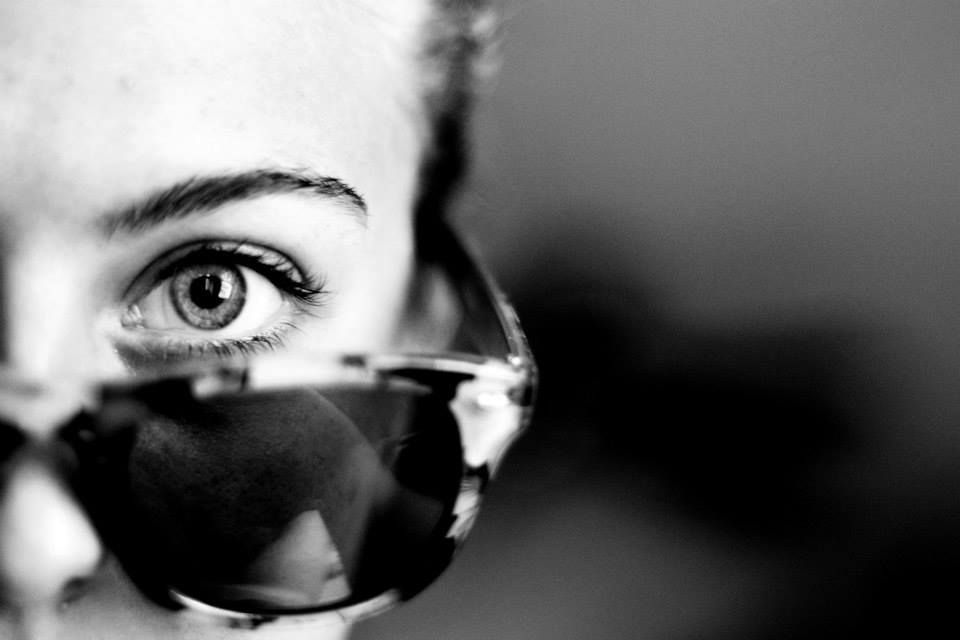 A close-up black and white photograph shows a dark sunglasses and an expressive eye peeking above the frame.