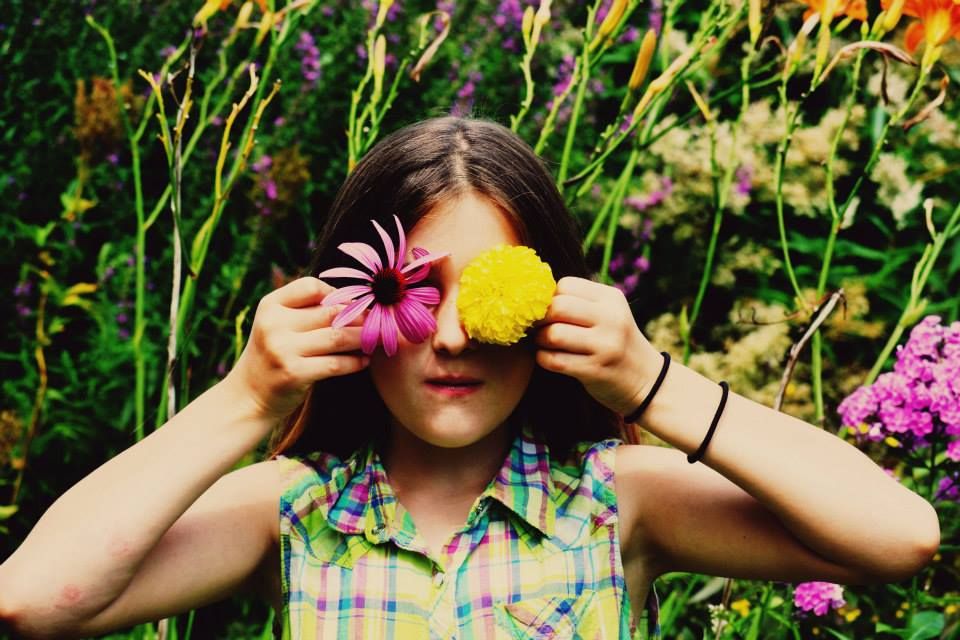 Someone holds colorful flowers over their eyes while posing in a vibrant garden with pink blooms in the background.