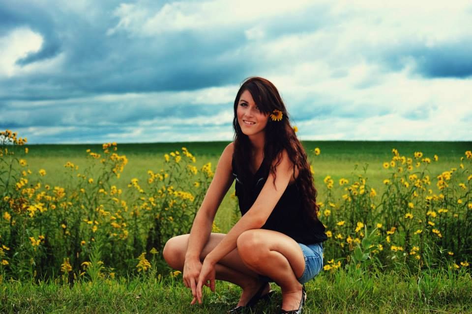 Woman crouching in a vibrant yellow canola field under dramatic cloudy skies on a summer day.