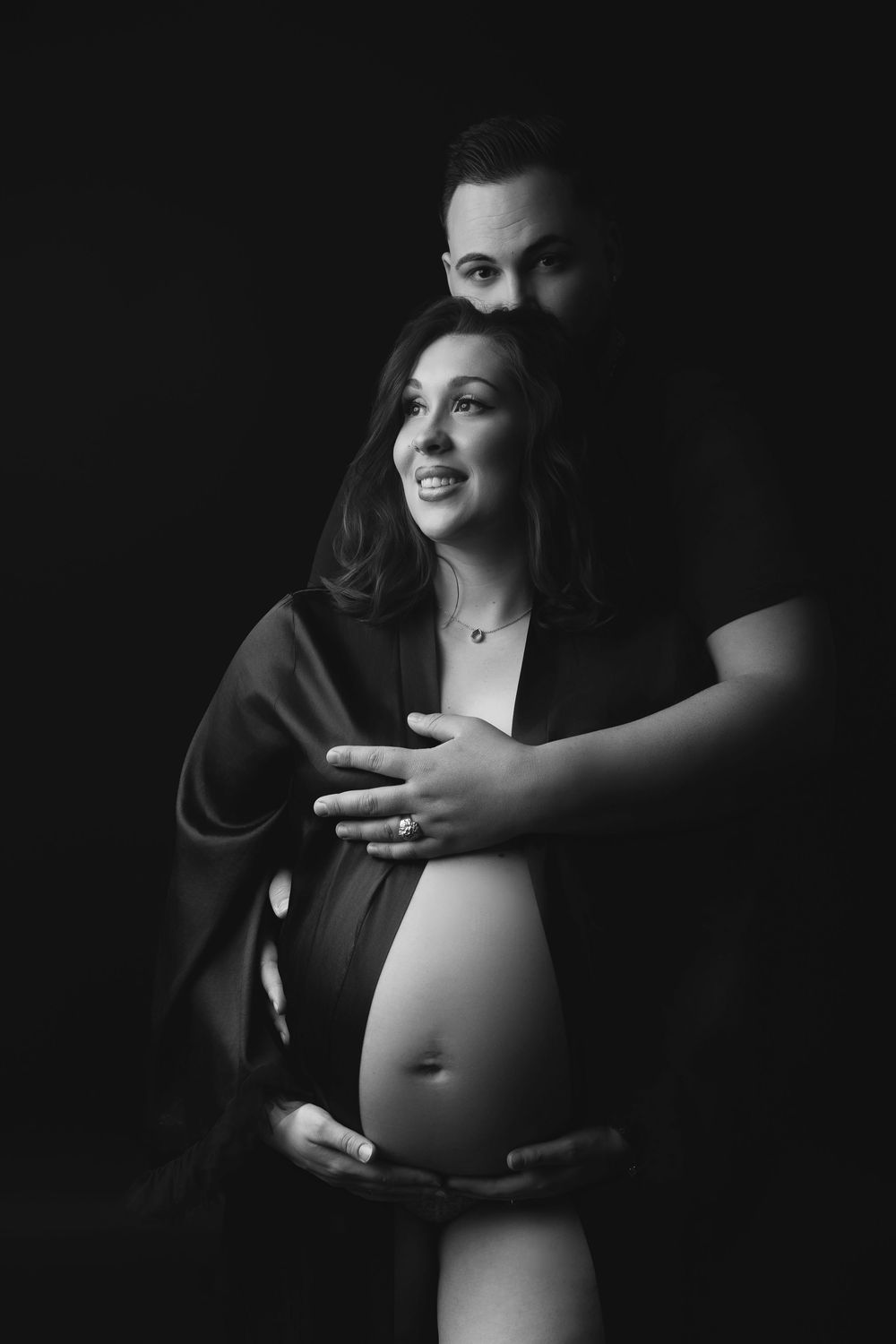 Black and white maternity photo showing a couple embracing, with the pregnant woman's bare belly visible.