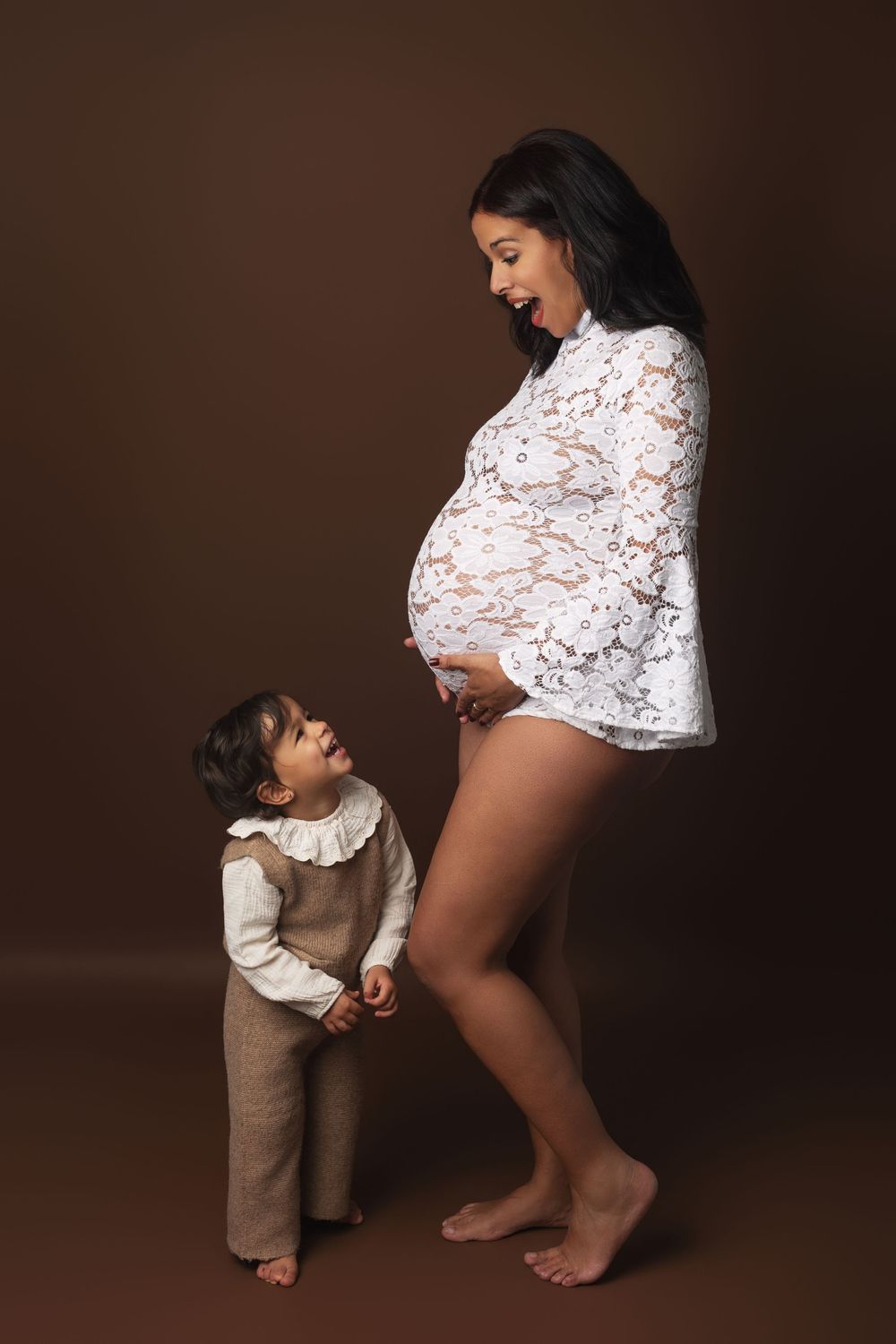 Pregnant woman in white lace top smiling at toddler looking up at her baby bump, maternity photo on brown background.
