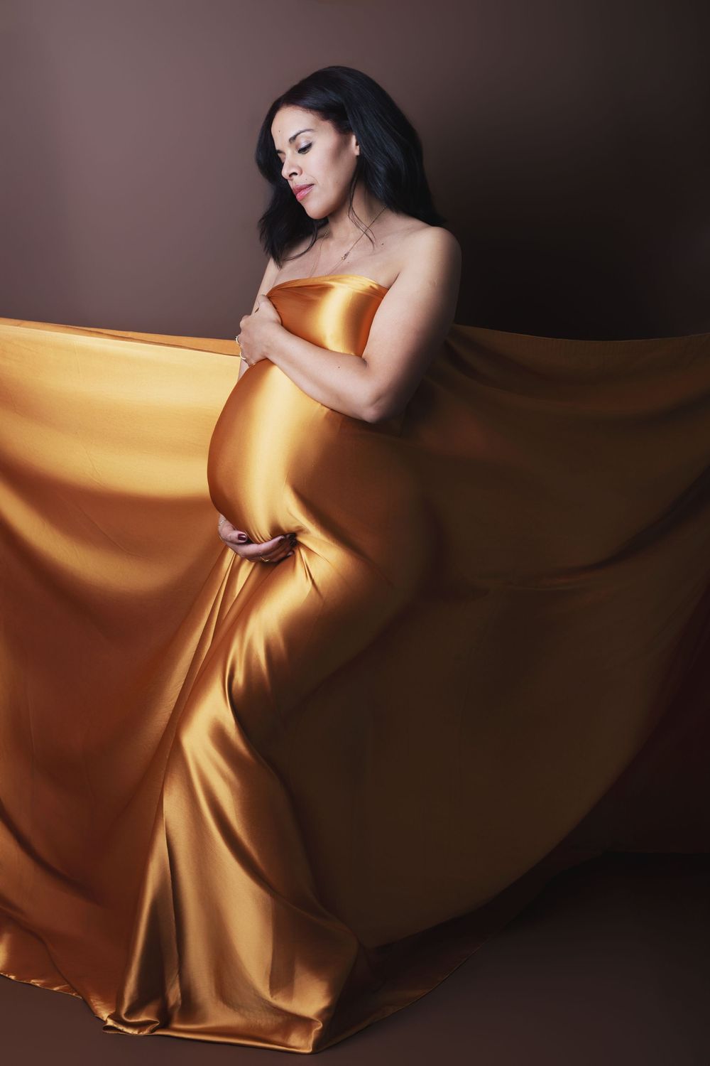 Pregnant woman draped in flowing golden satin fabric, cradling her baby bump against a dark brown studio background.