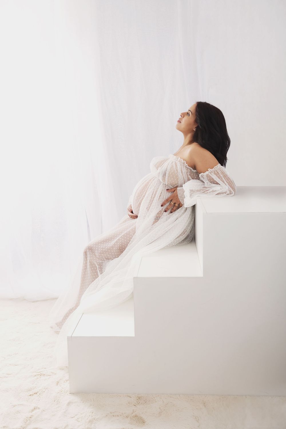 Pregnant woman in white lace gown sitting on white steps, cradling baby bump in bright airy studio setting.