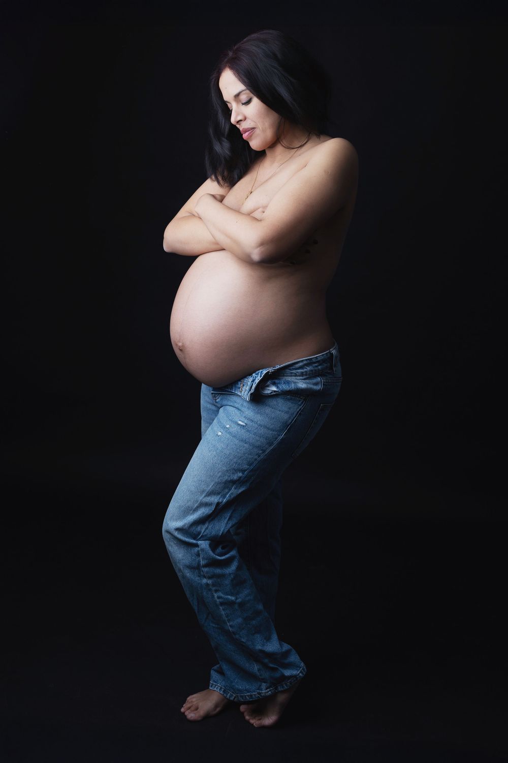 Pregnant woman in blue jeans with arms crossed, posing against black background in artistic maternity photo.