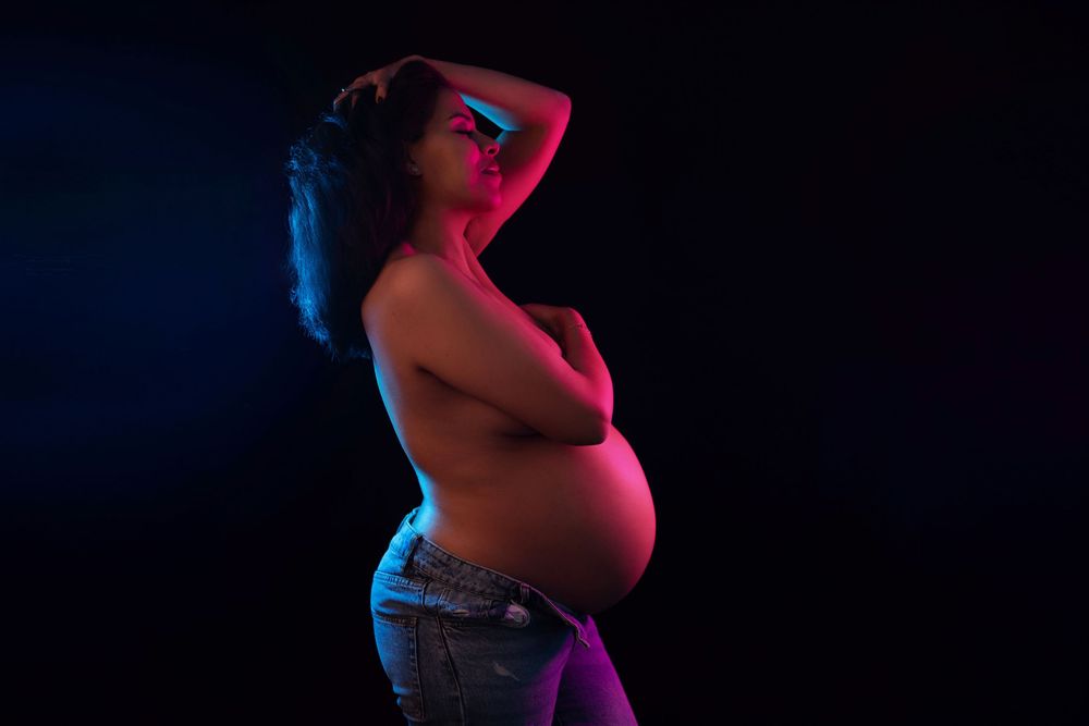 Pregnant woman posing topless in dramatic neon pink and blue studio lighting against dark background.