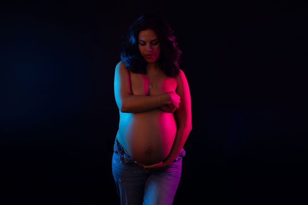 Pregnant woman in dramatic pink and blue neon lighting cradling her bare baby bump against dark background.