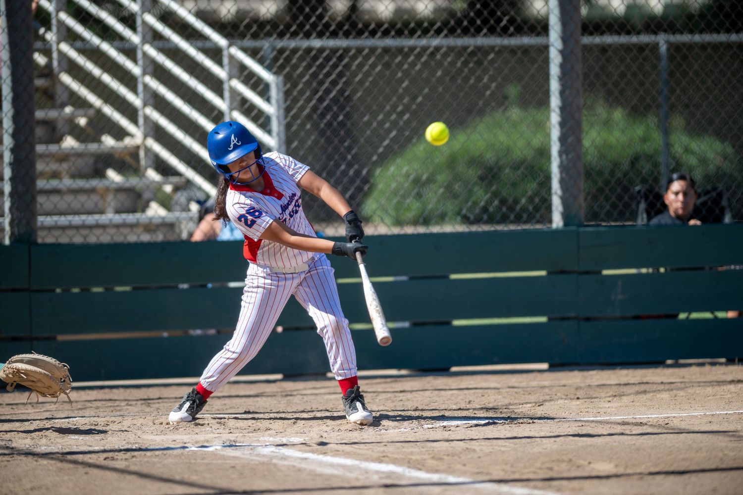 Female softball player in red and white uniform swinging bat at yellow ball during competitive game.