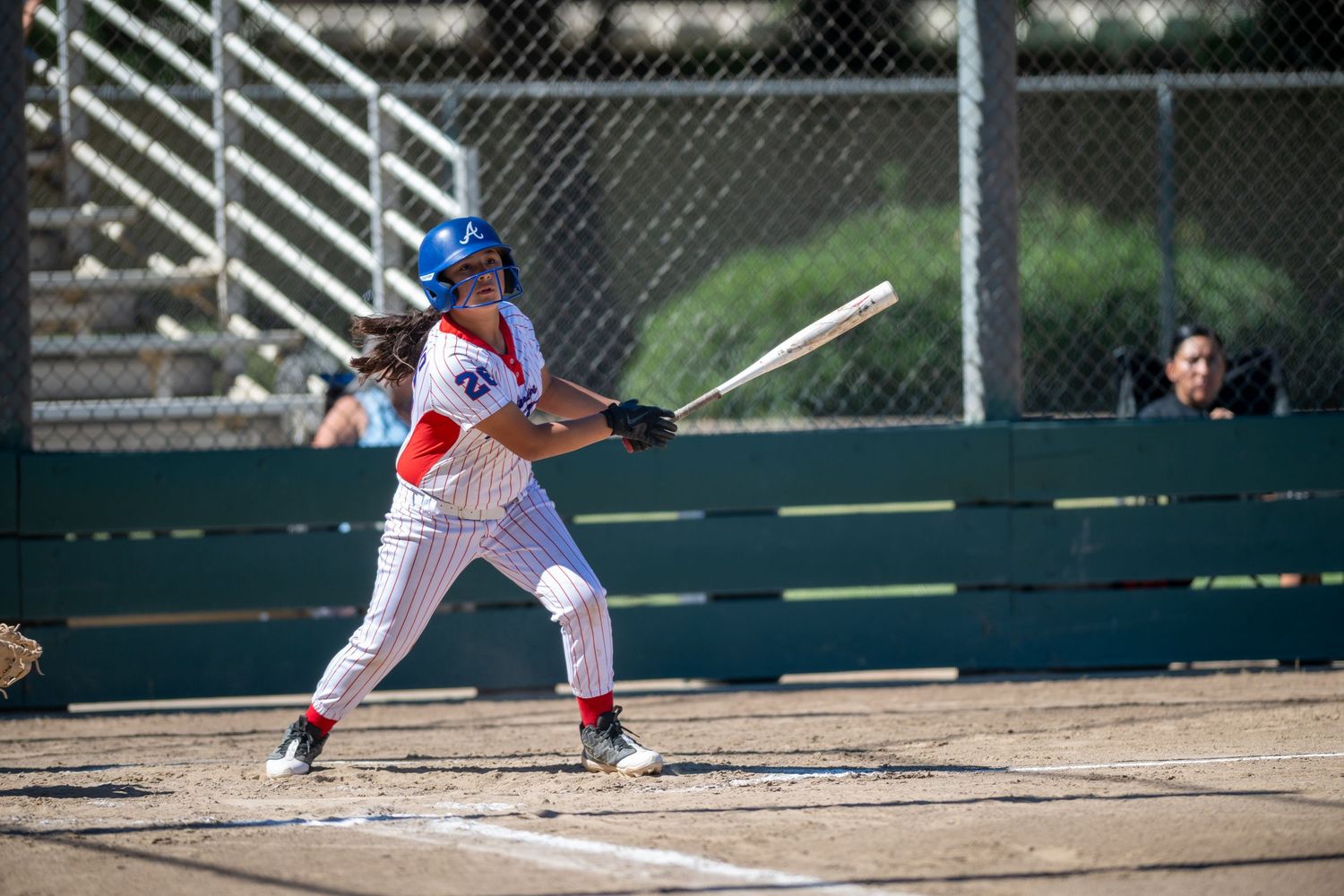 Female baseball player in red pinstripe uniform swinging bat during a sunny outdoor game.