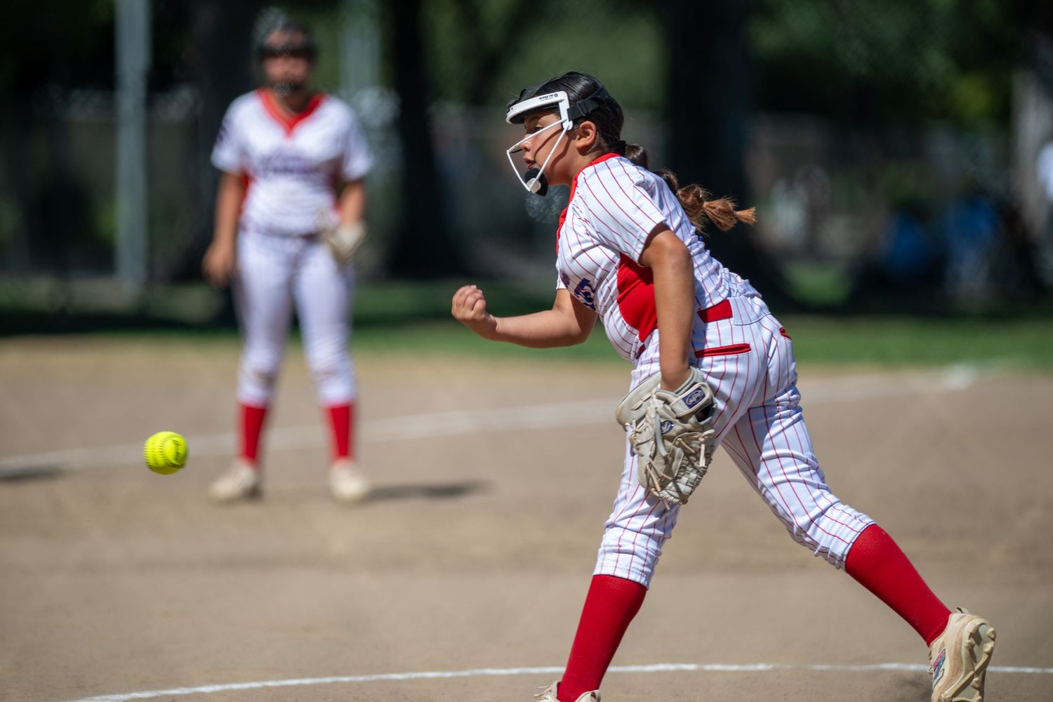 Softball pitcher in red and white uniform mid-throw during a sunny outdoor game, with teammate visible in background.