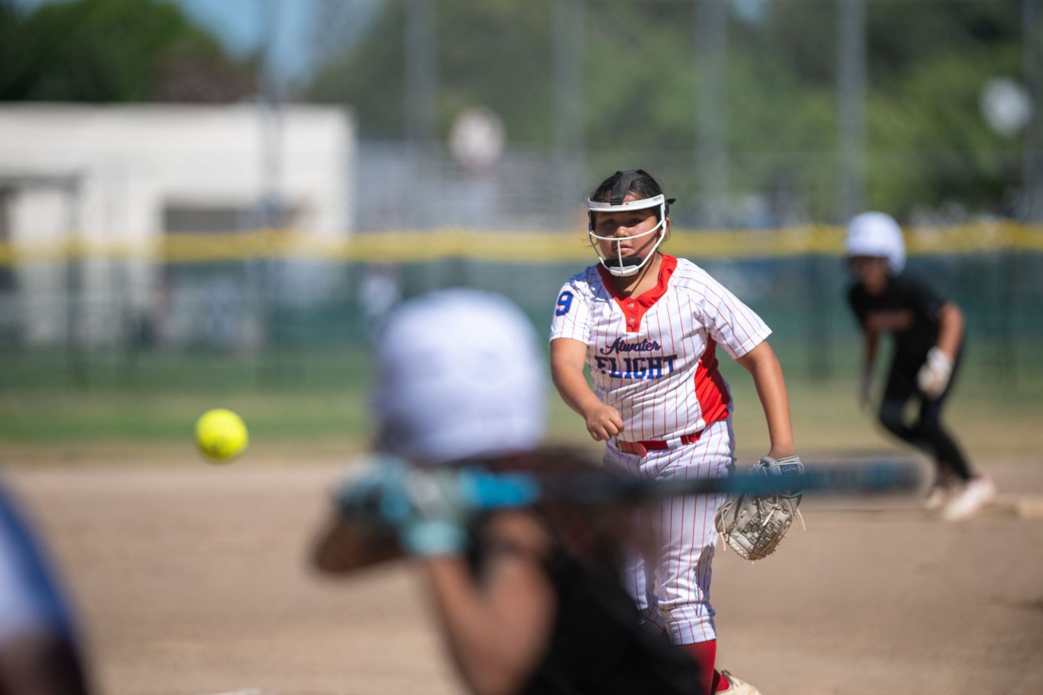 Softball pitcher in red uniform delivers a pitch during a sunny youth softball game on a green field.
