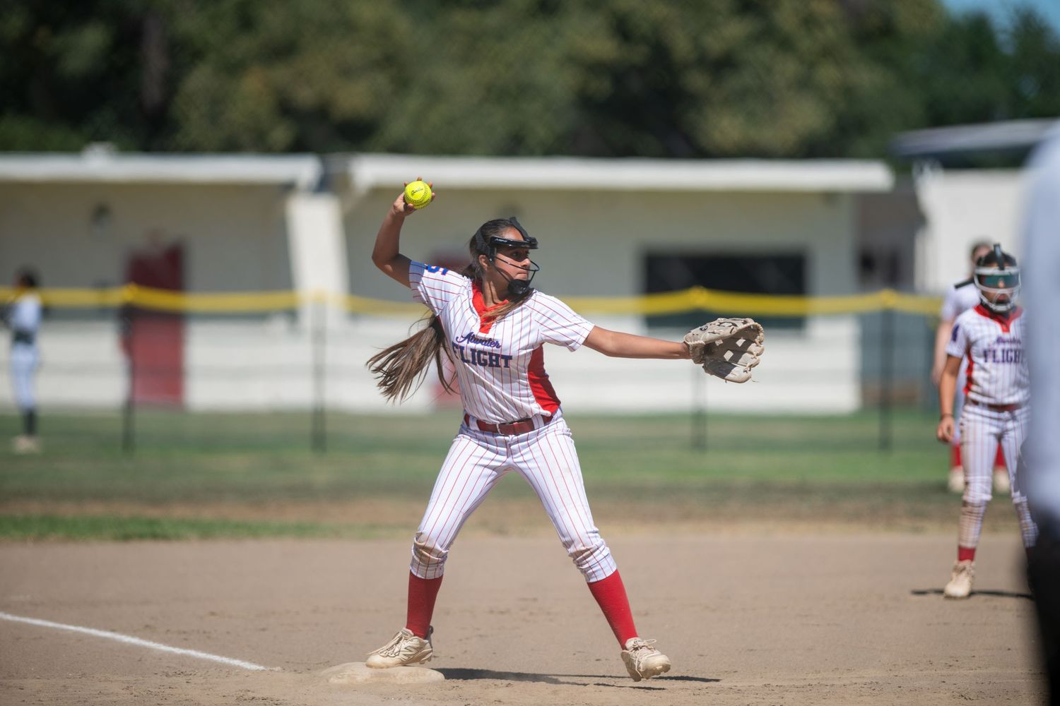 Female softball player in red and white uniform winds up to throw the ball during a sunny outdoor game.