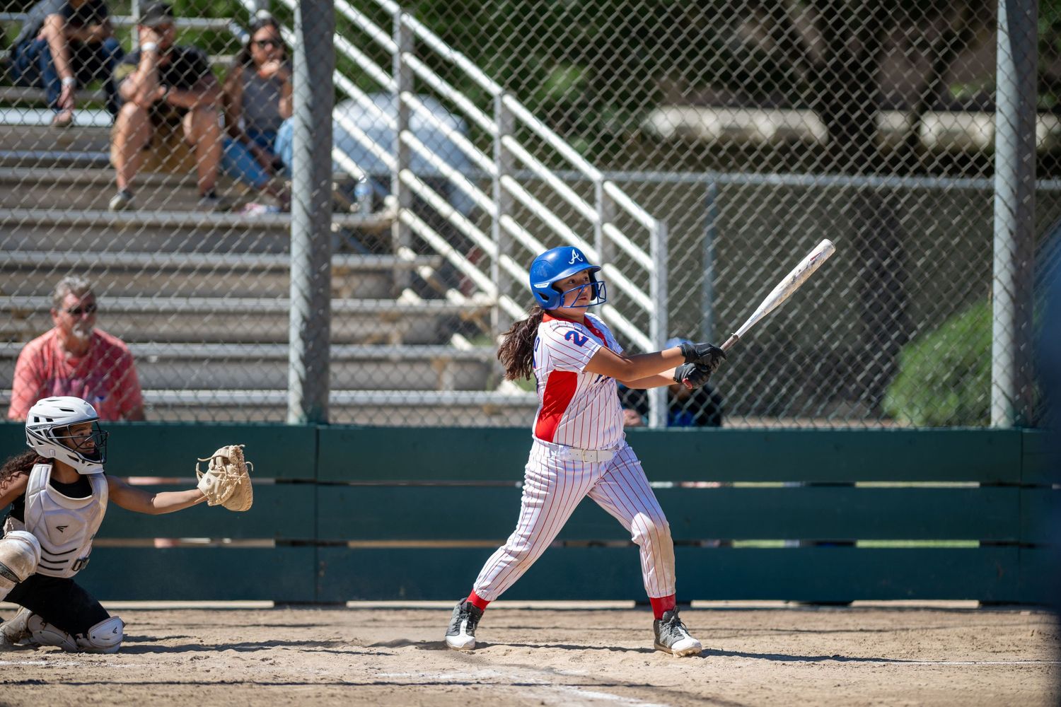Youth baseball batter in red, white, and blue uniform swings at pitch during sunny outdoor game.