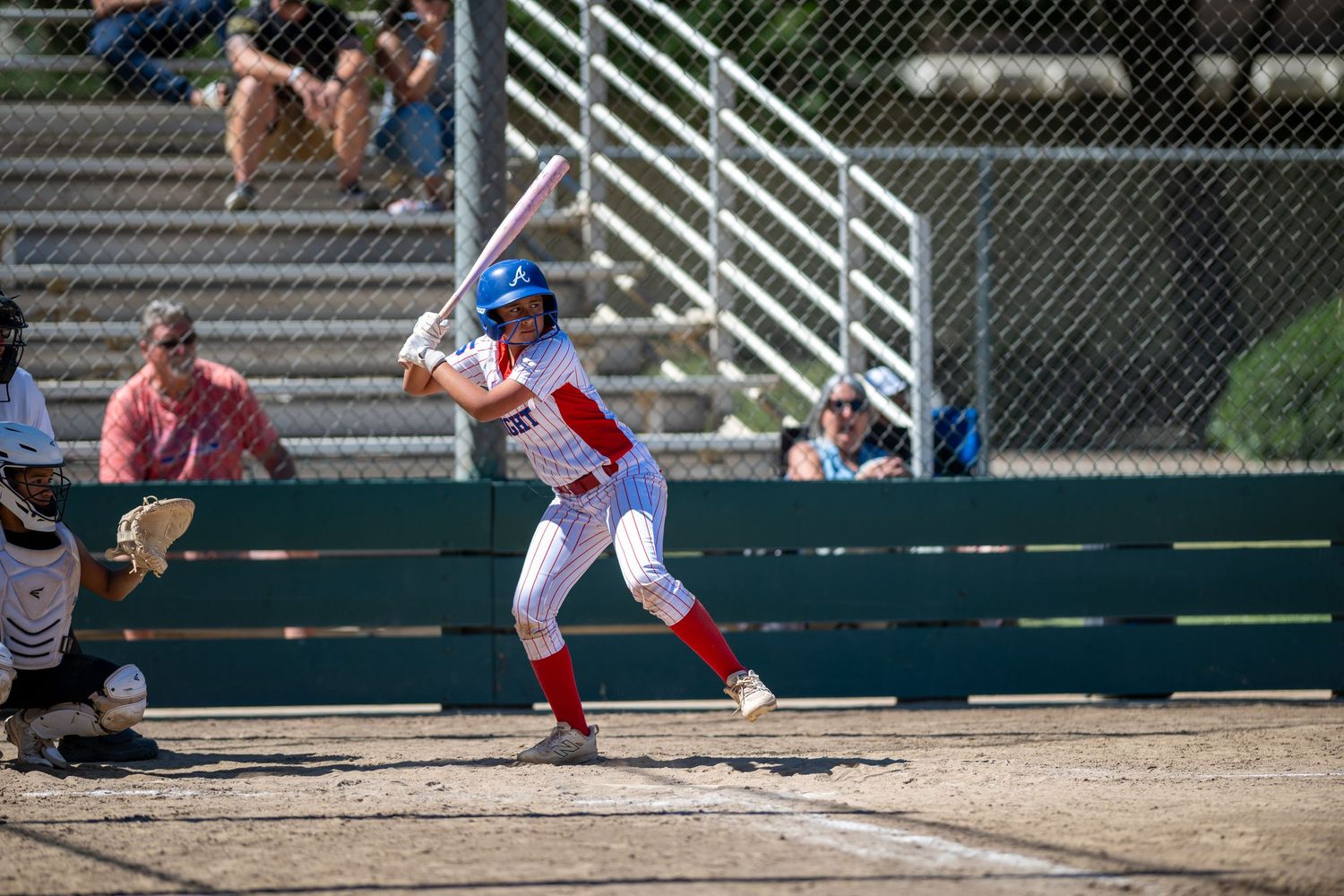 Female softball batter in red and white uniform with blue helmet stands ready at plate during sunny outdoor game.