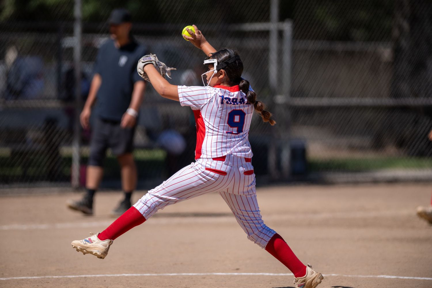 Softball pitcher #9 in red and white pinstripe uniform winds up to throw during a sunny outdoor game.