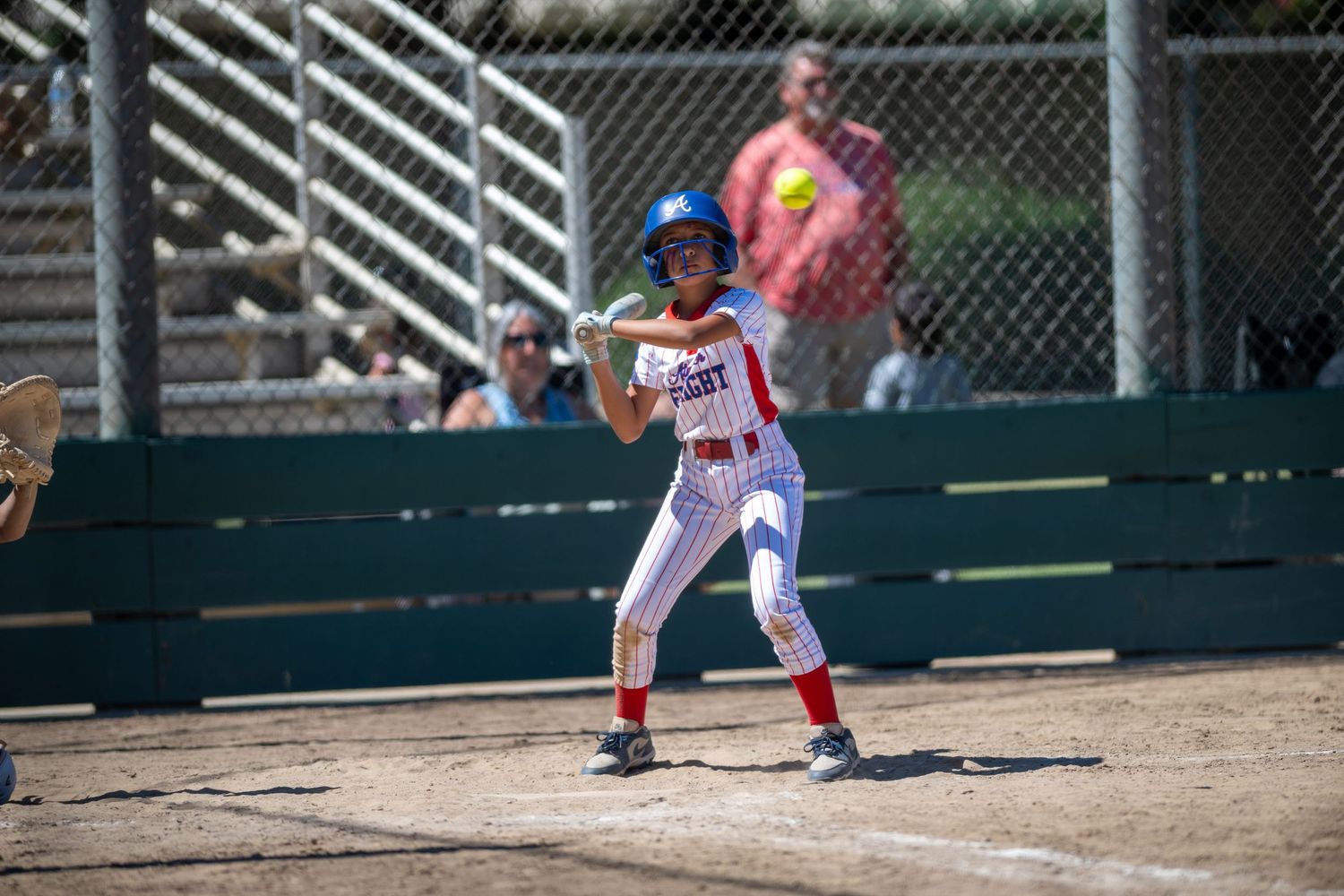 Female softball batter in red and white uniform with blue helmet stands ready at home plate on a sunny day.