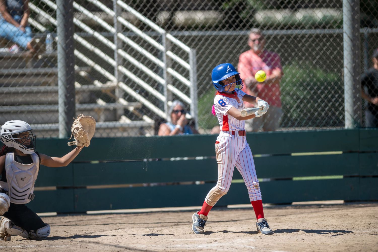 Softball batter in red and white uniform swings at pitch during competitive game, catcher ready behind home plate.