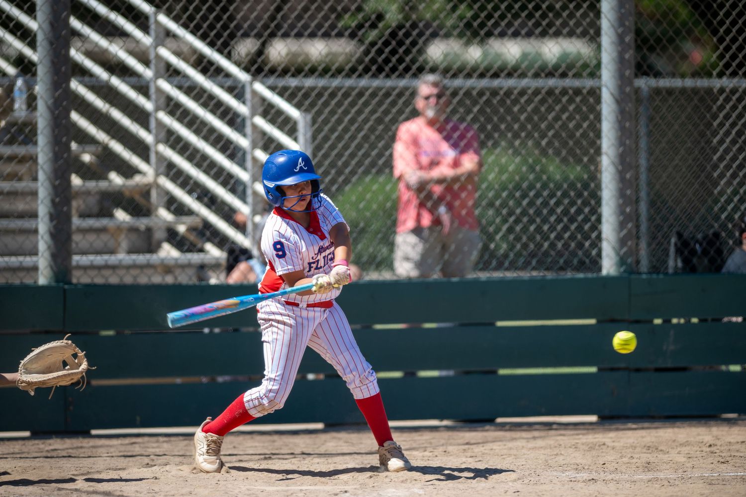 A softball player in a red and white uniform swings a blue bat at a yellow ball during a sunny outdoor game.