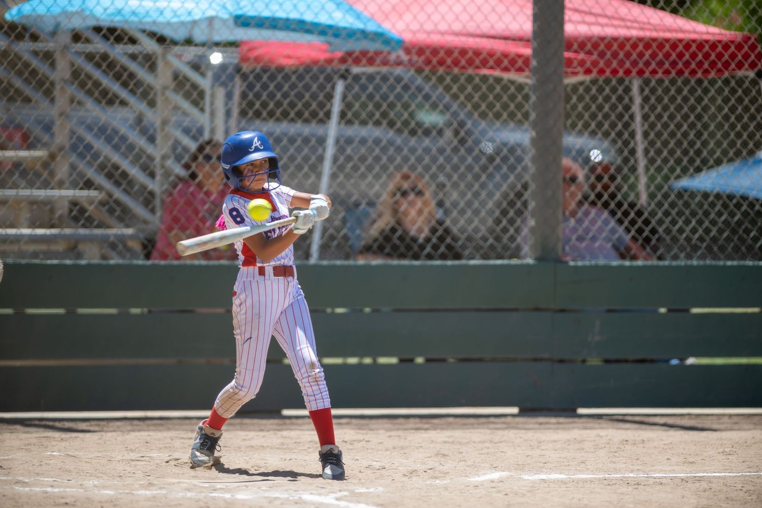 Female softball player in red and white pinstripe uniform swinging bat during a sunny outdoor game.