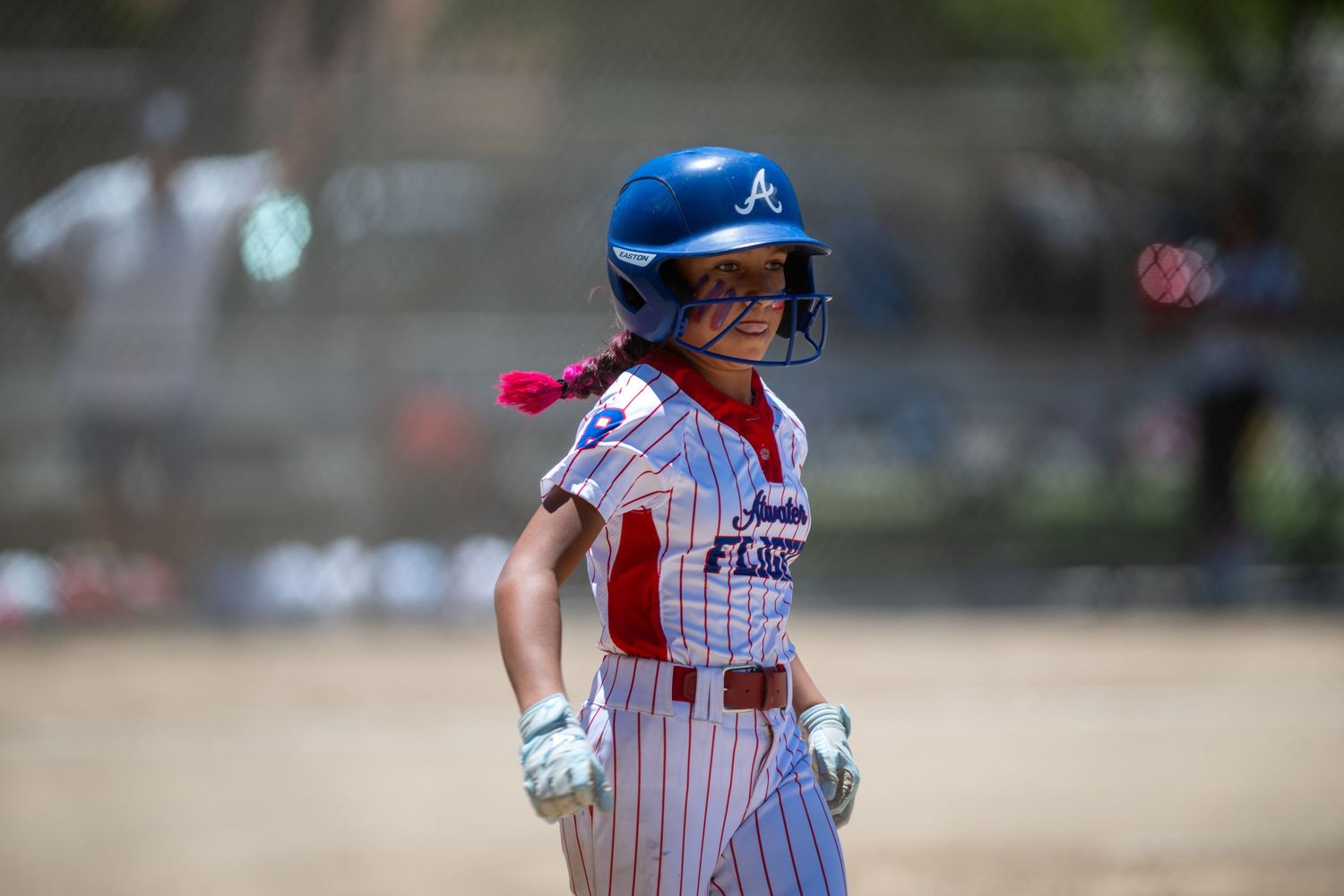 Female softball player in blue helmet and white uniform stands on base during a sunny outdoor game.