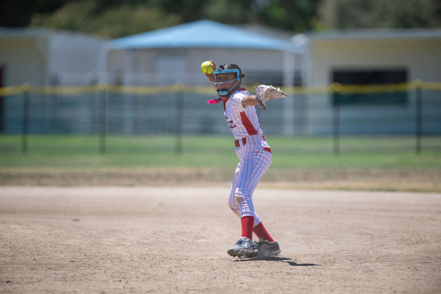 Softball player in red and white uniform throws ball during an outdoor game on a dirt infield.