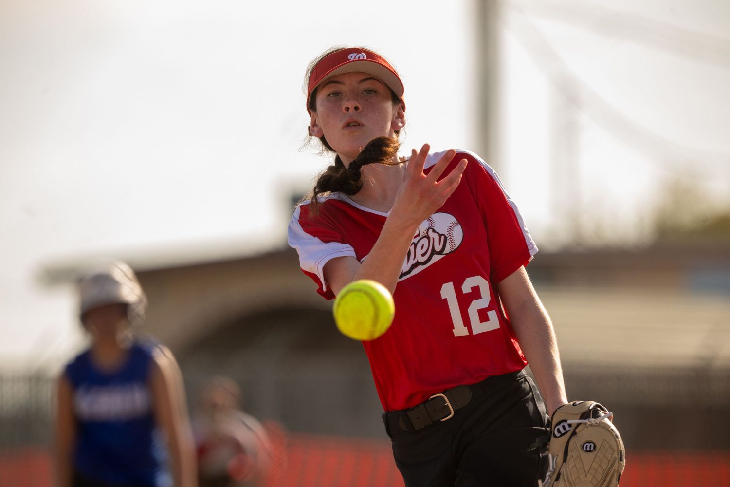 A softball pitcher in a red uniform throws a yellow ball during a game.