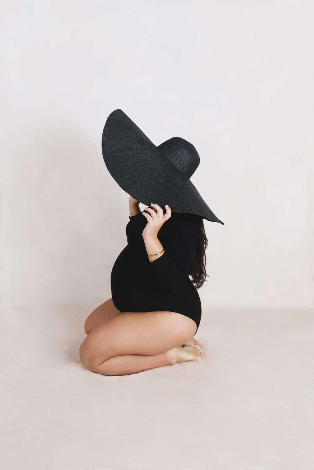 Minimalist maternity photos with a dramatic black sun hat against white background.