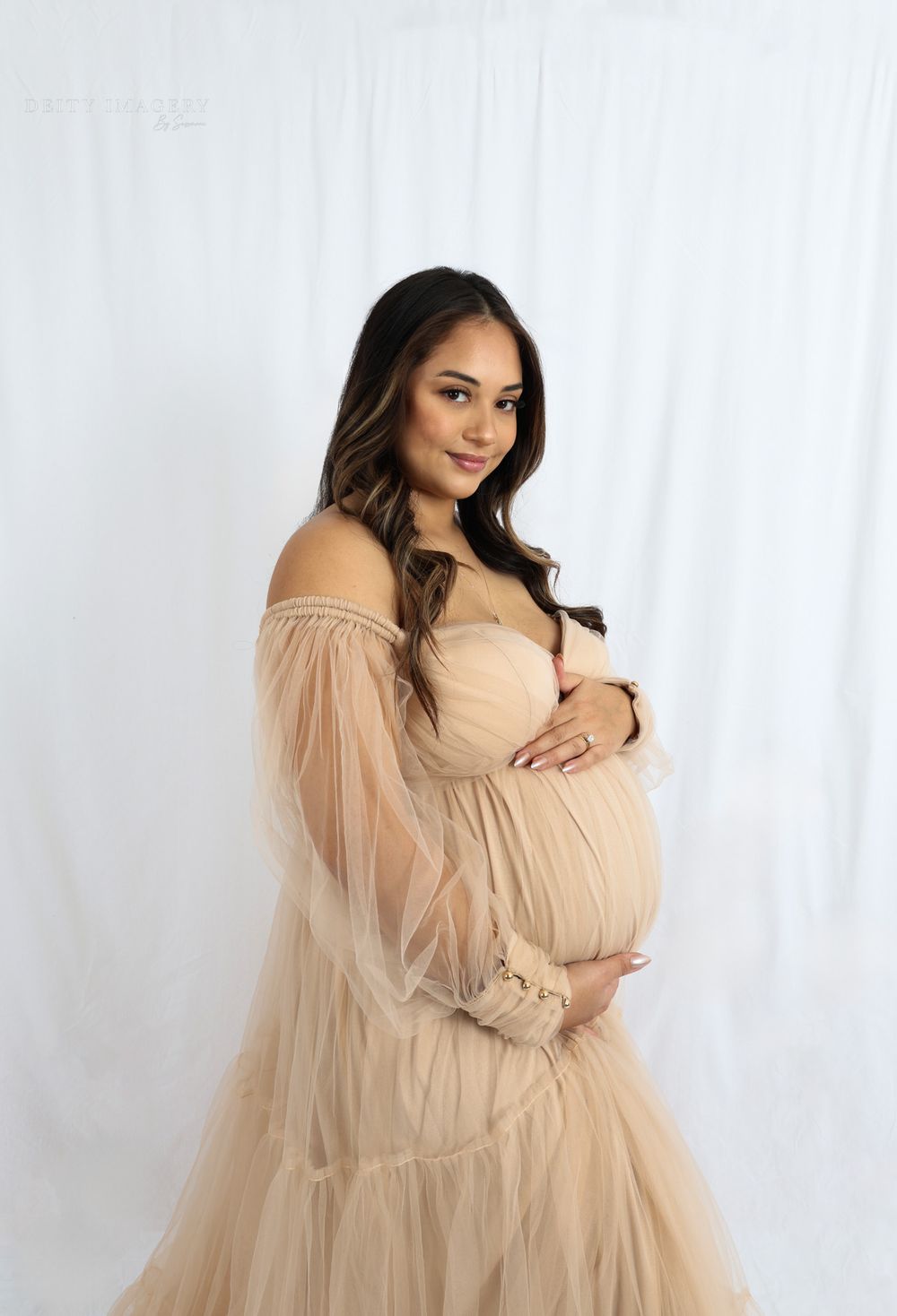 Ethereal maternity photoshoot featuring a flowing champagne tulle gown against a white backdrop.