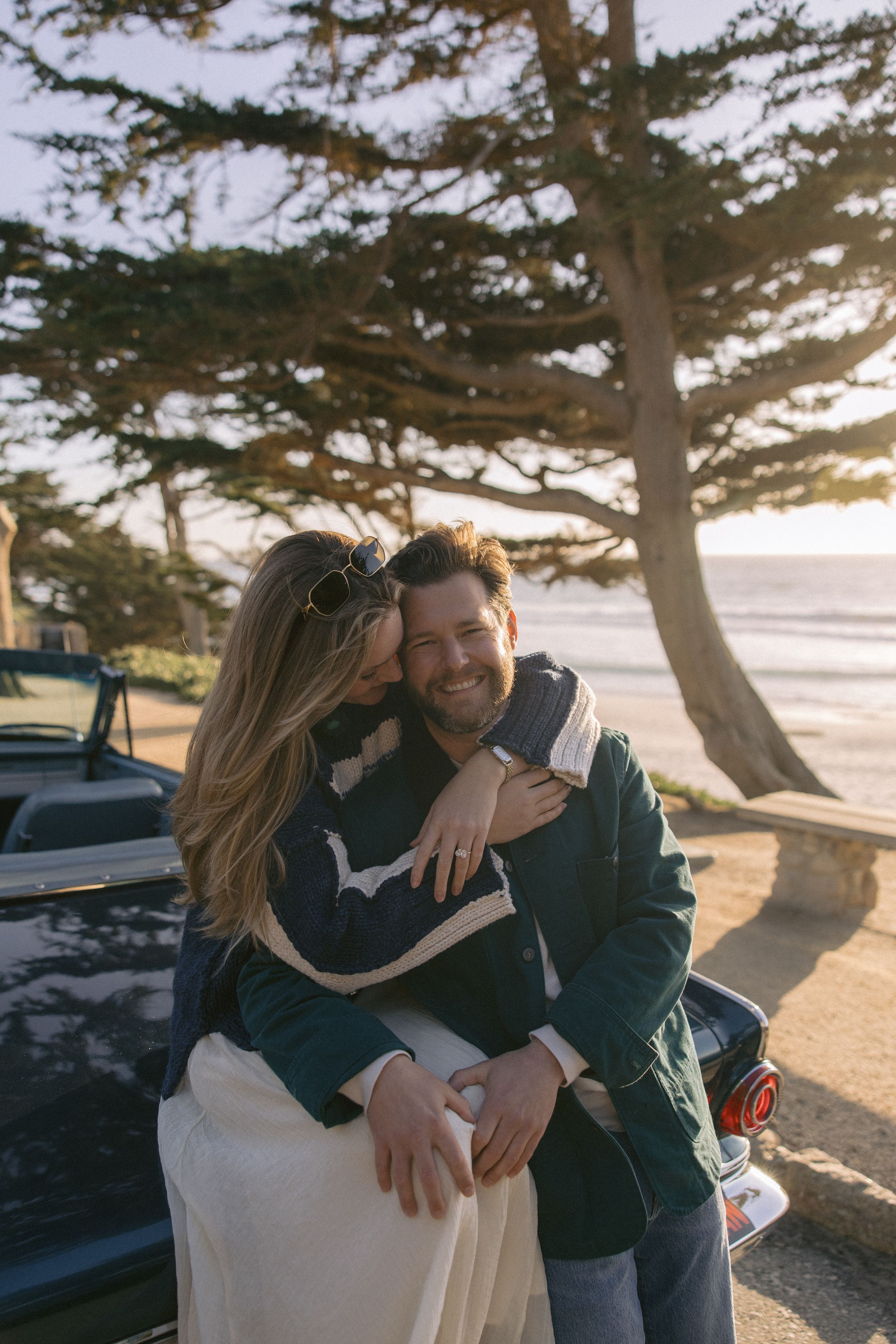 Couple embracing on California coast at sunset with cypress tree and ocean view in background.