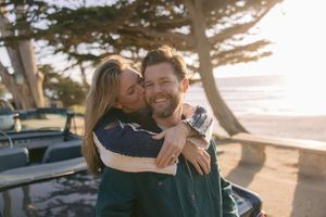A couple embraces and smiles together at sunset by the beach with cypress trees in the background.