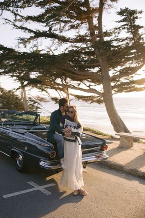 A couple embraces next to a vintage car overlooking the ocean with cypress trees framing the sunset scene.