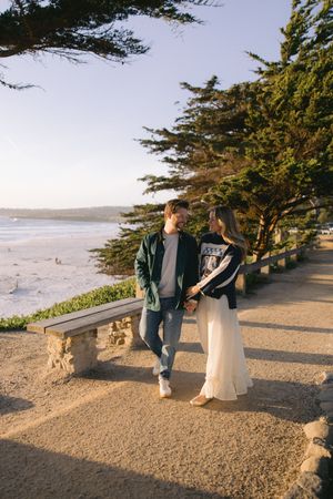 A couple walks along a scenic coastal path with cypress trees and ocean views during golden hour at sunset.