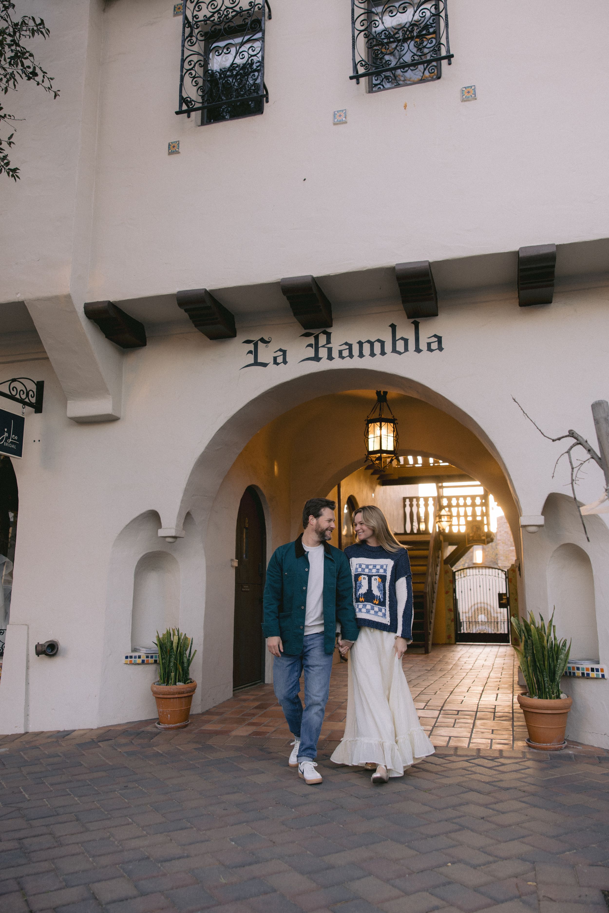 A Spanish-style arched entryway reading 'La Rambla' with potted plants and decorative wood beams overhead.