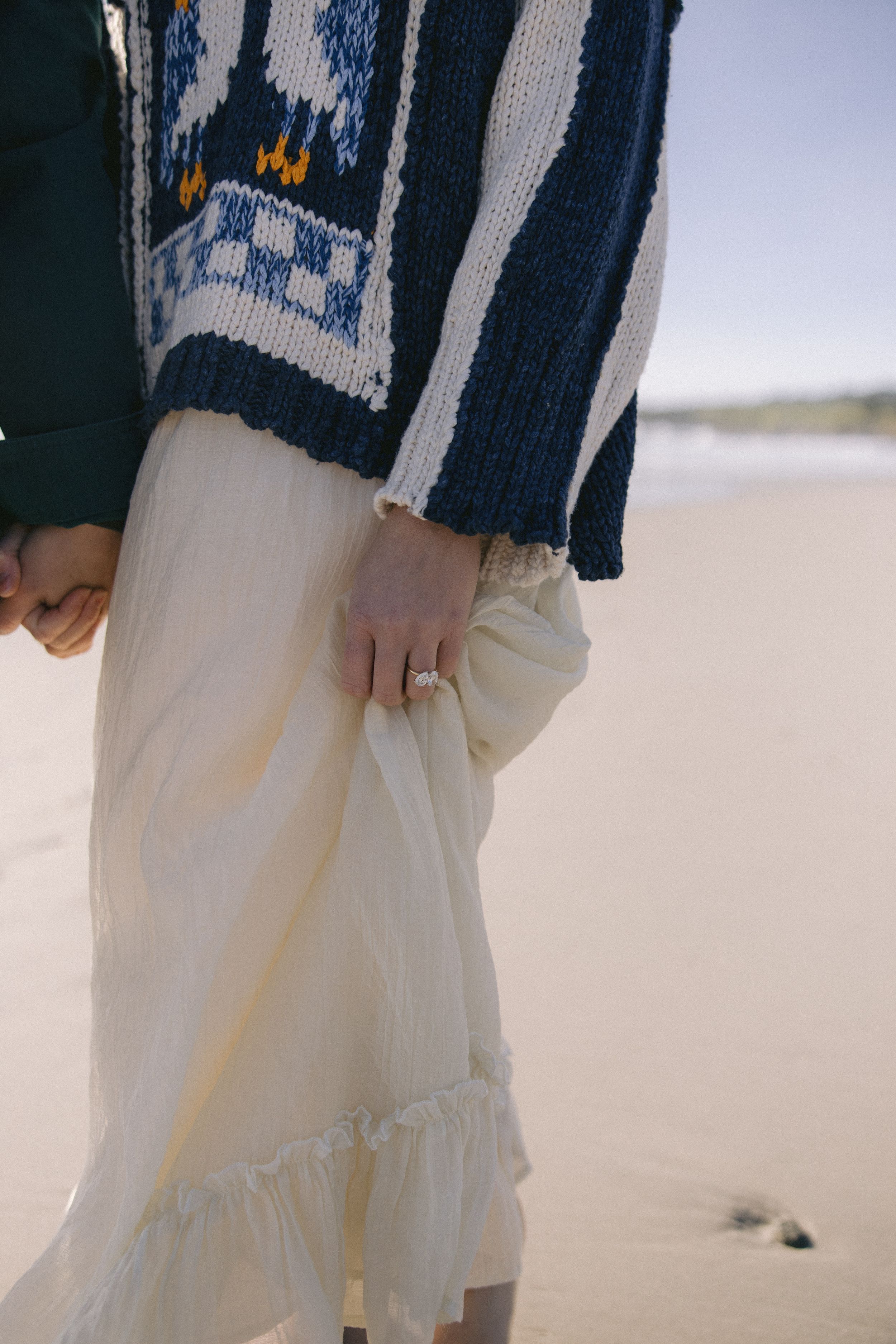 Someone wearing a navy patterned knit sweater and cream ruffled maxi skirt stands on a sandy beach.
