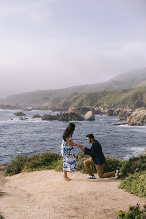 A couple shares an intimate proposal moment on a cliff overlooking the rugged Big Sur coastline on a misty day.
