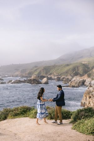 Two people standing on a coastal cliff path overlooking rugged rocky shores and crashing waves along Big Sur.