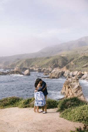 Scenic coastal view at Big Sur with two people embracing on clifftop trail overlooking rocky shoreline and mountains.