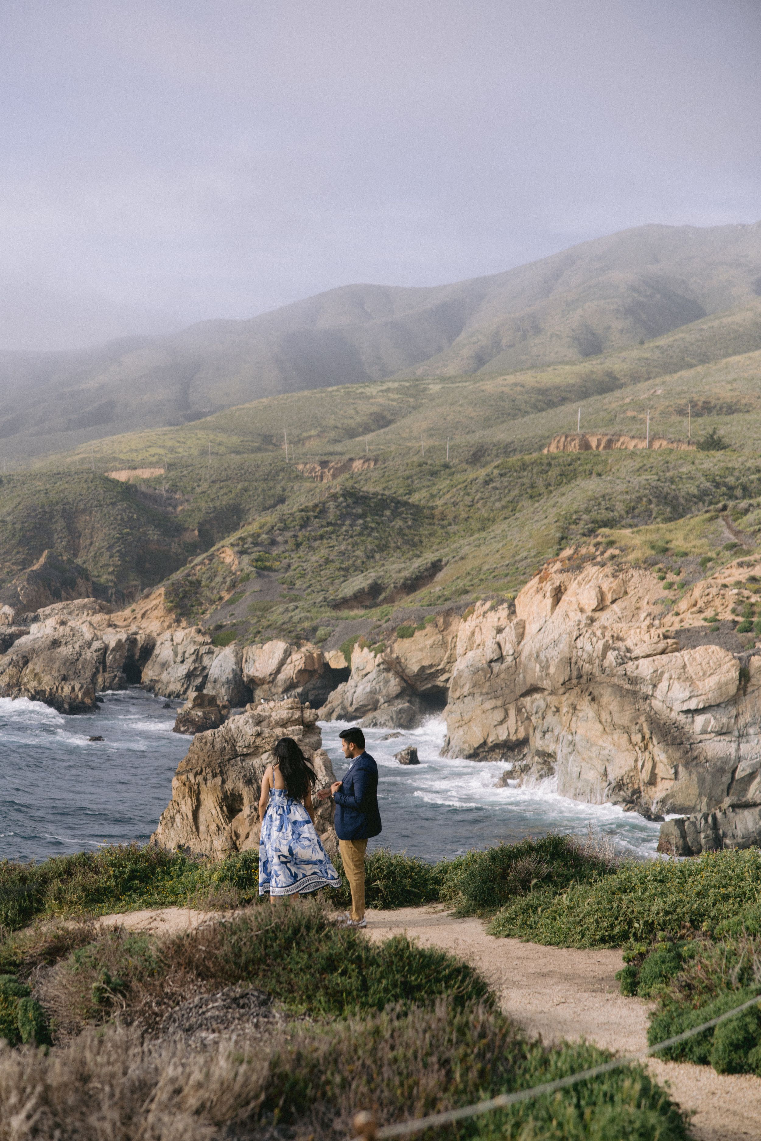 Two people stand on a coastal trail overlooking rugged cliffs and crashing waves along the Pacific coast on a foggy day.