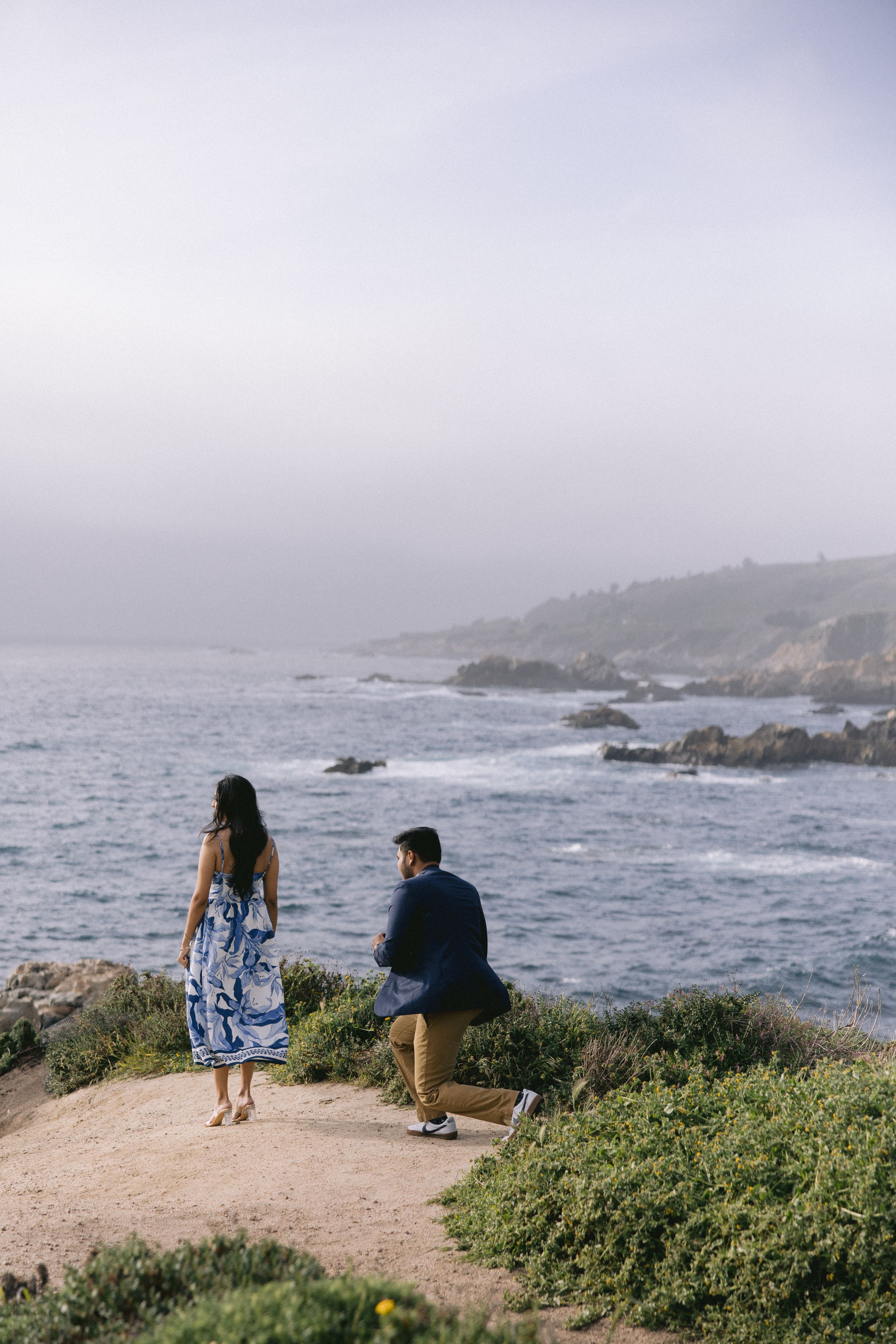 Two people stand on a coastal path overlooking rocky Pacific Ocean shores on a misty afternoon.