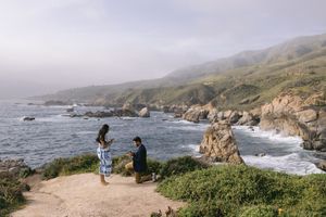 Two people enjoying scenic views along the rugged Big Sur coastline with rocky cliffs and crashing waves.