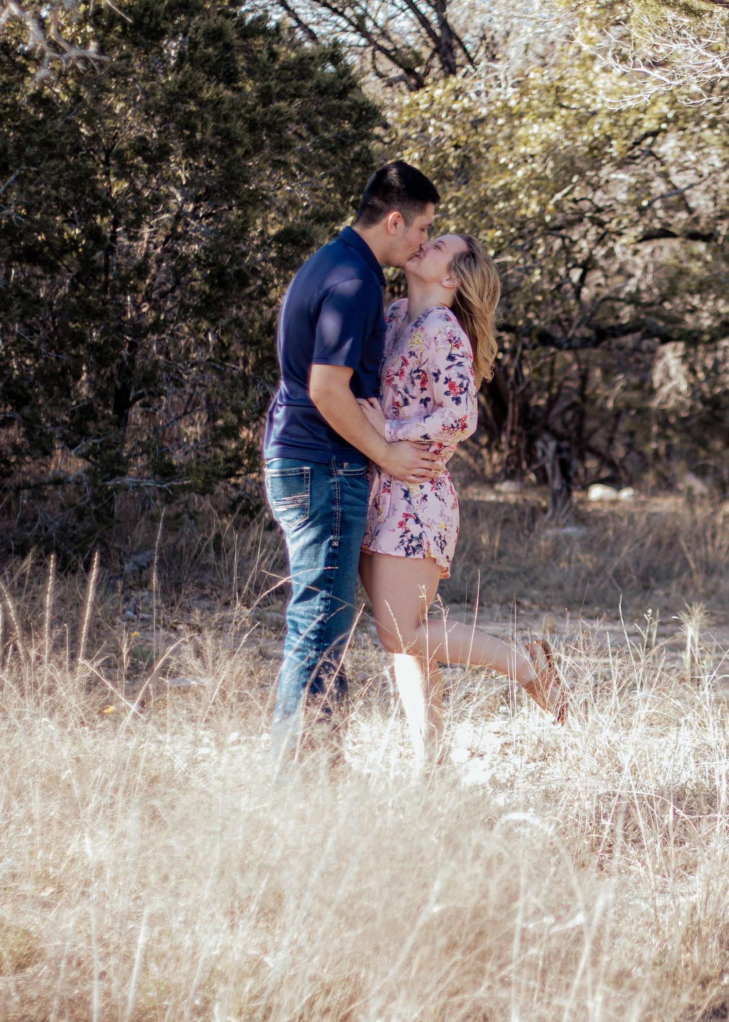 A couple shares a romantic moment outdoors in sunlight, with her wearing a floral top and him in a navy shirt.