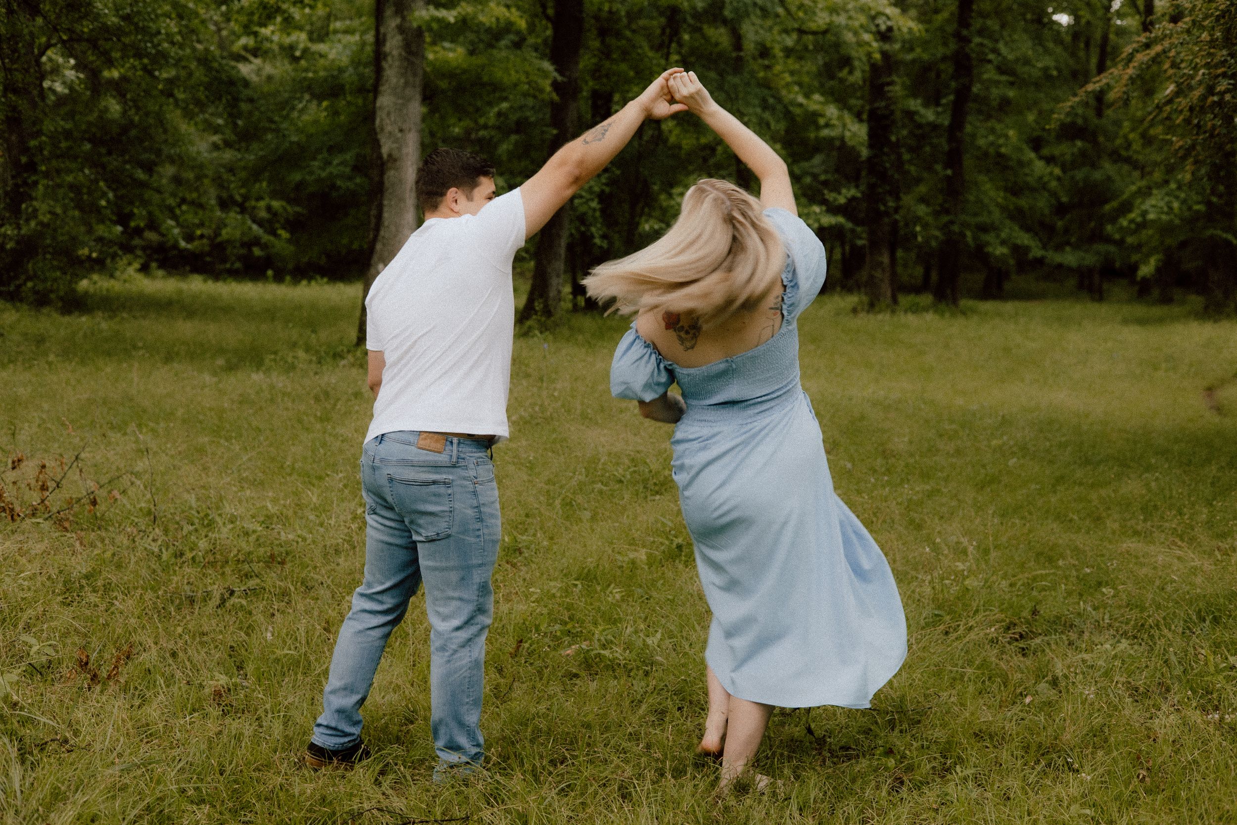 Two people in casual clothing dancing together in a grassy field surrounded by trees on a sunny day.