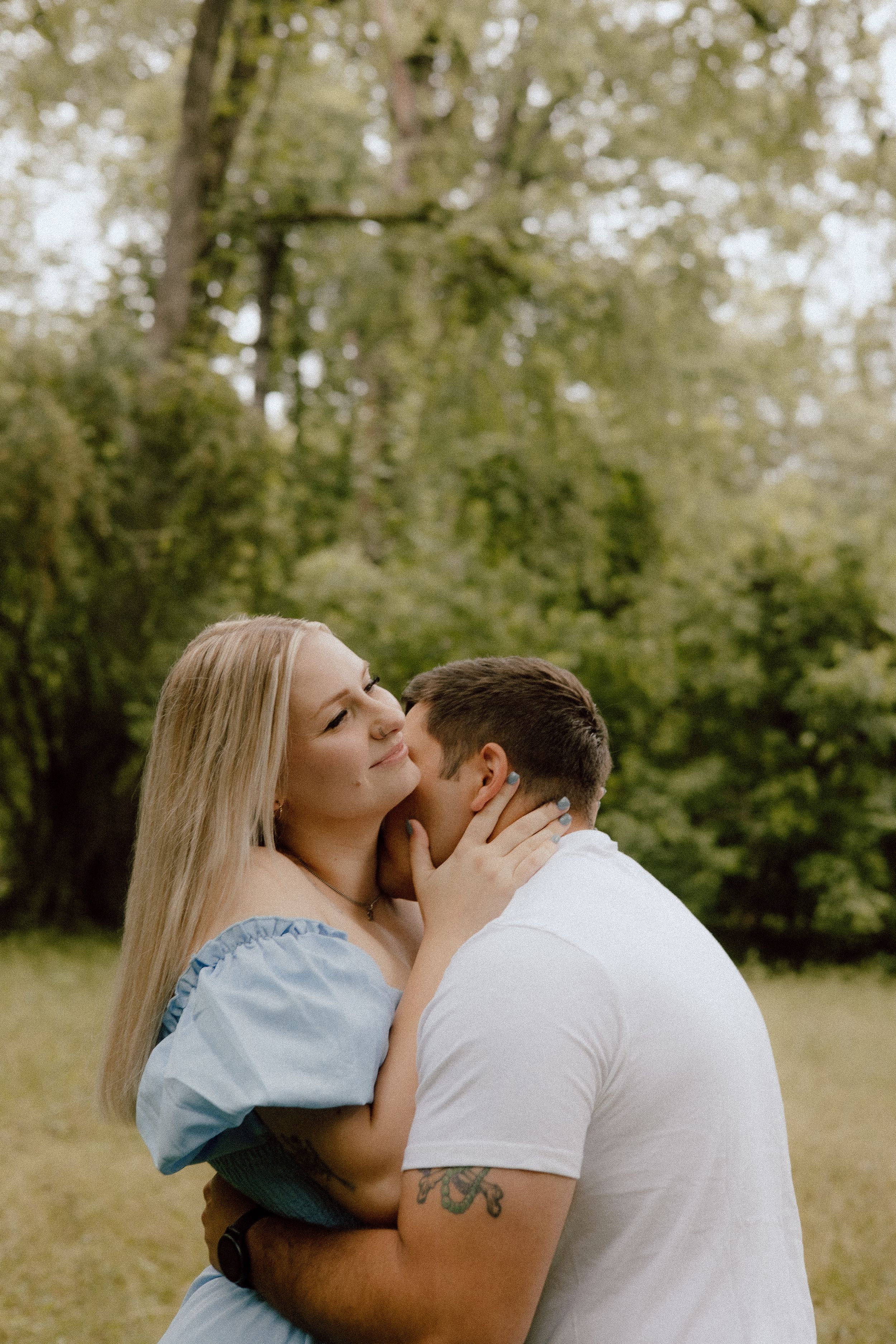 A romantic couple share an intimate moment outdoors in a natural setting among trees and greenery.