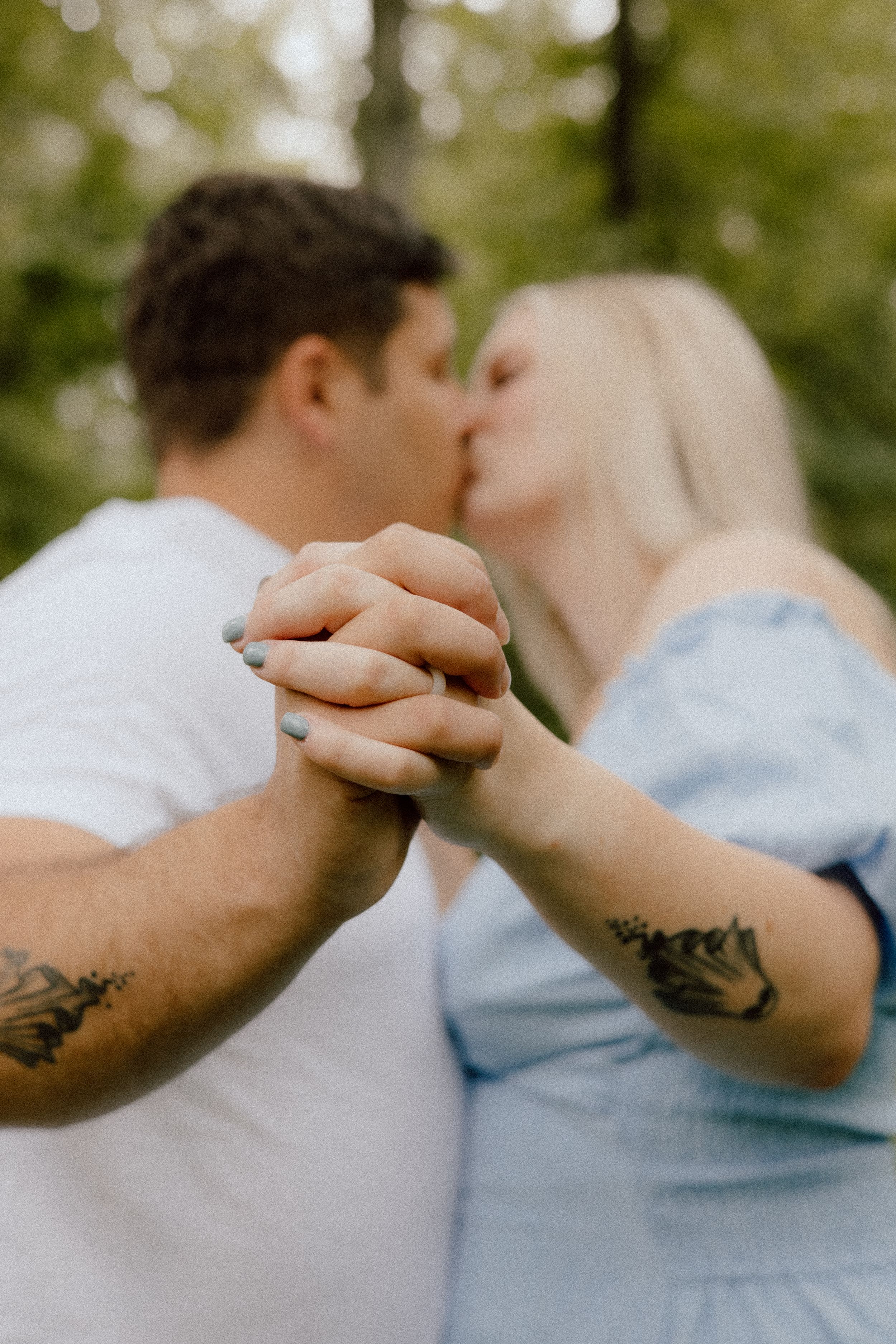 A couple holding hands and kissing while showing their arm tattoos in a romantic outdoor setting.