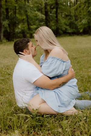 A couple sits intimately together on grassy ground in a wooded area, with the woman wearing a light blue off-shoulder dress.